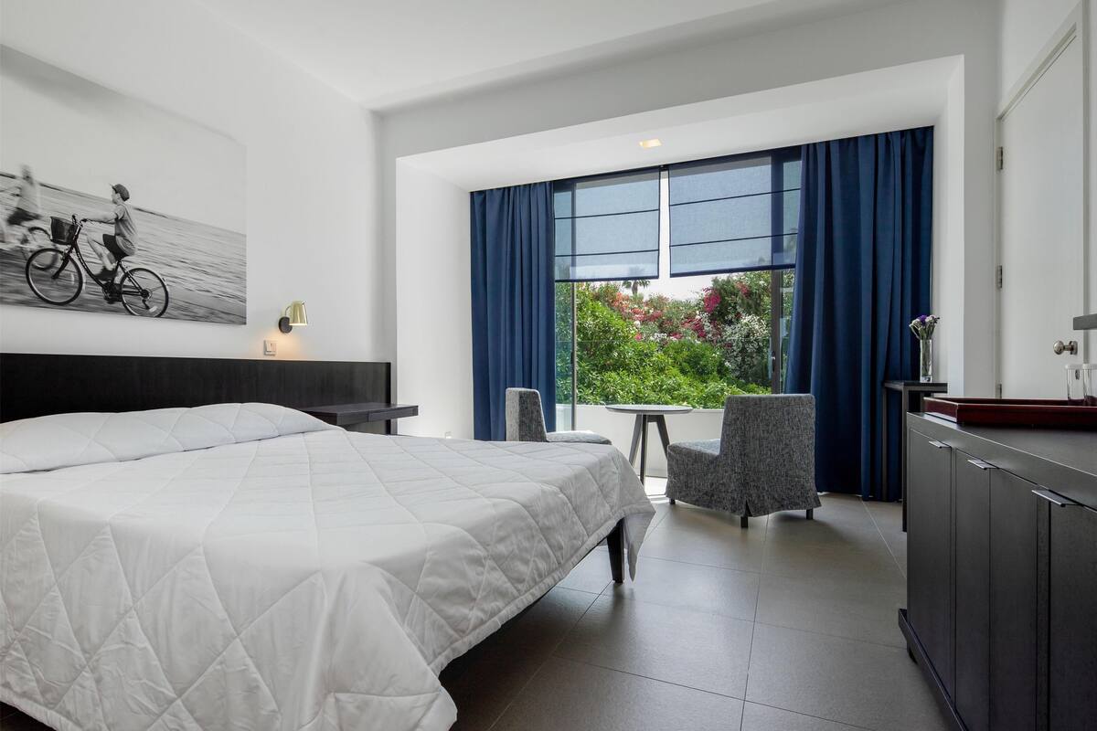 A hotel room with a large bed, a desk, a chair, a small table, and a view of greenery through a window.