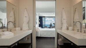 Une salle de bain moderne dans un hôtel, dotée de deux lavabos, d’un grand miroir et offrant une vue sur une chambre avec un lit et un balcon donnant sur la mer.