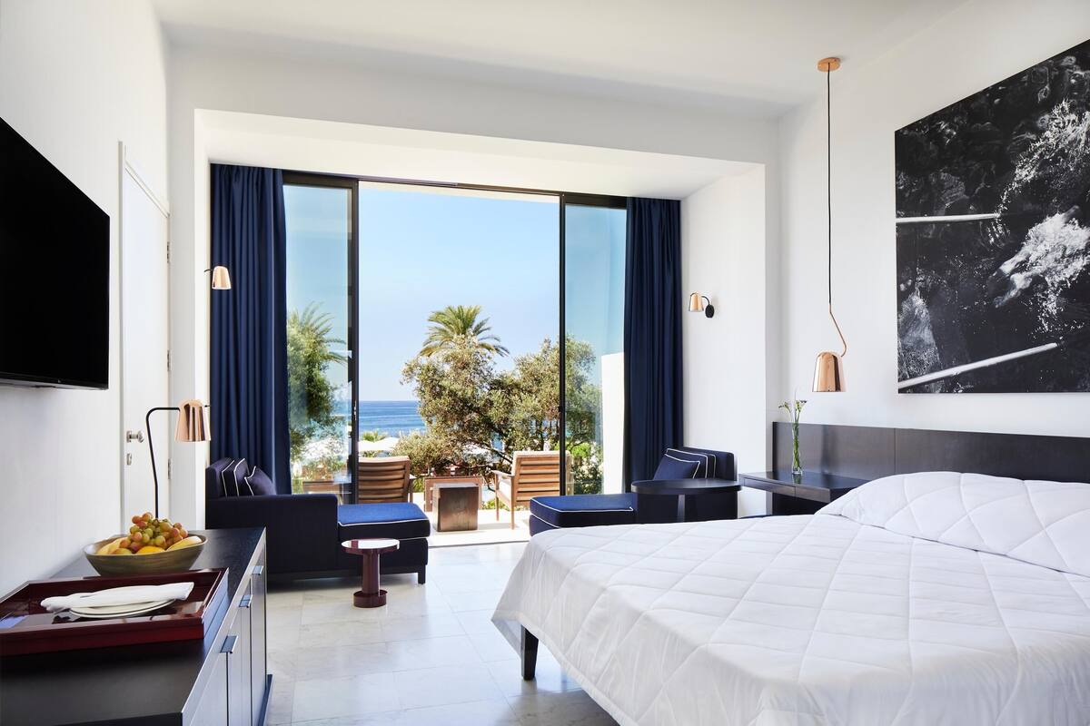 A modern hotel room with a large bed, a TV, a dining area with a fruit bowl, and a balcony with a view of the ocean.