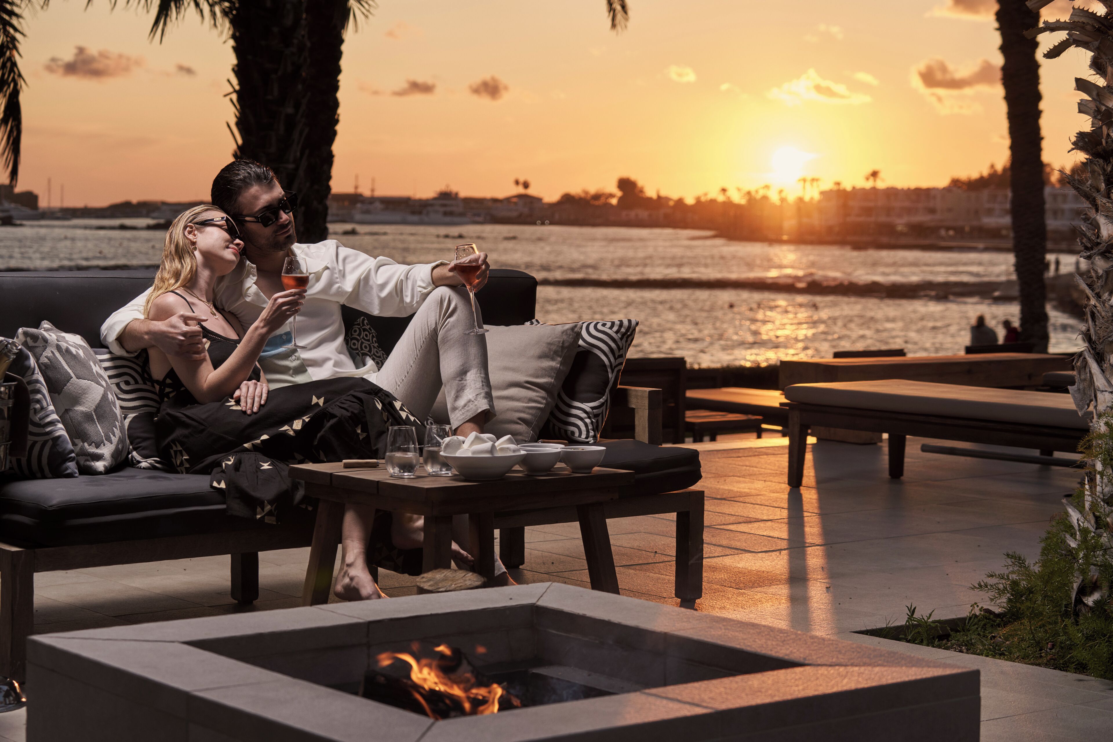 A couple relaxing on a sofa with a view of the sunset over the water, a fire pit, and outdoor seating.
