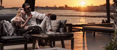A couple relaxing on a sofa with a view of the sunset over the water, a fire pit, and outdoor seating.