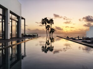 A modern infinity pool with a view of the sunset and palm trees.