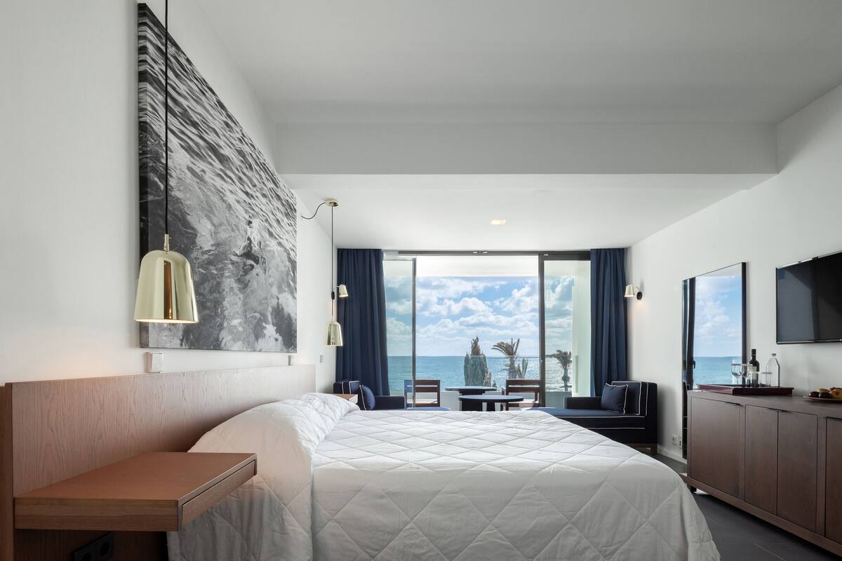 A modern hotel room with a large bed, a desk, a dining area, and a view of the sea.