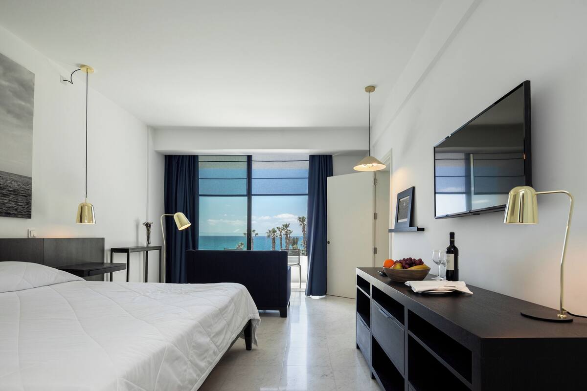 A modern hotel room with a bed, bedside table, desk, and a large window offering a view of the ocean.