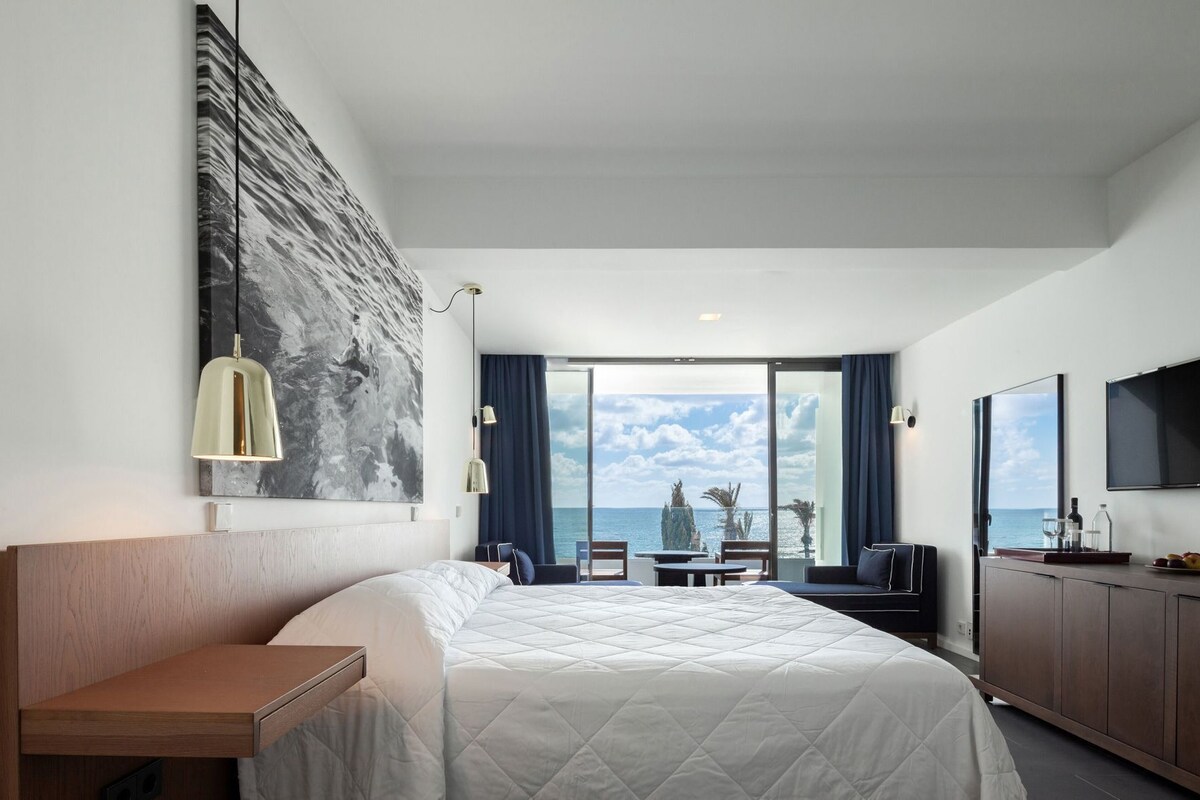 Habitació d'hotel amb un llit, un escriptori, un televisor, una finestra gran que dona al mar i un quadre al mur.