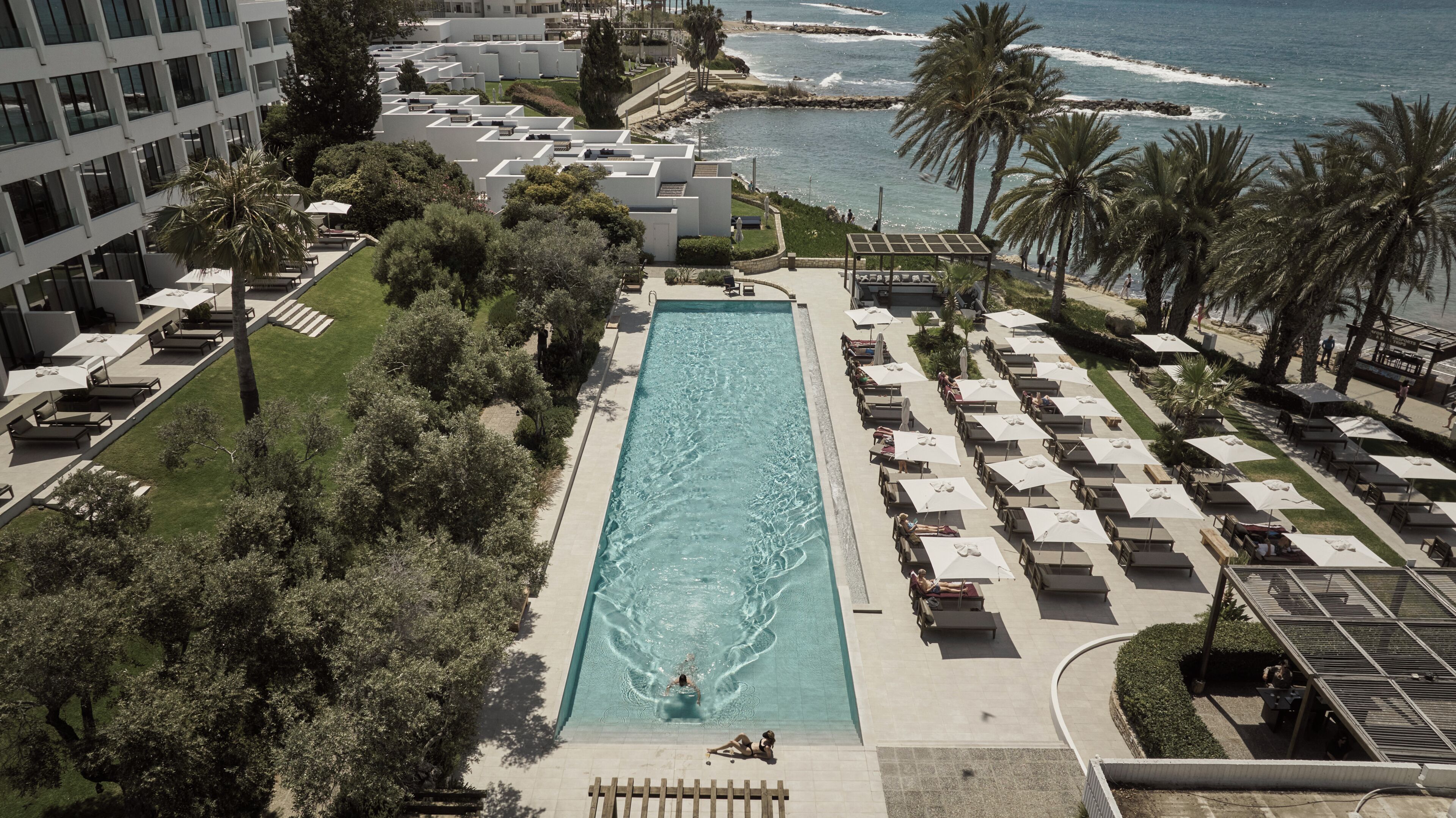 A pool area with lounge chairs and umbrellas, surrounded by greenery and a view of the ocean.
