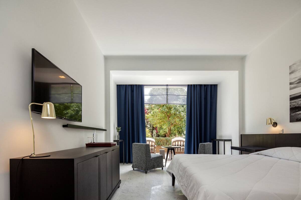 A modern hotel room with a bed, a desk, a chair, a TV, and a view of the outdoors.