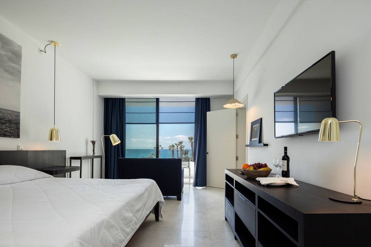 A modern hotel room with a bed, bedside table, desk, and a large window offering a view of the sea.