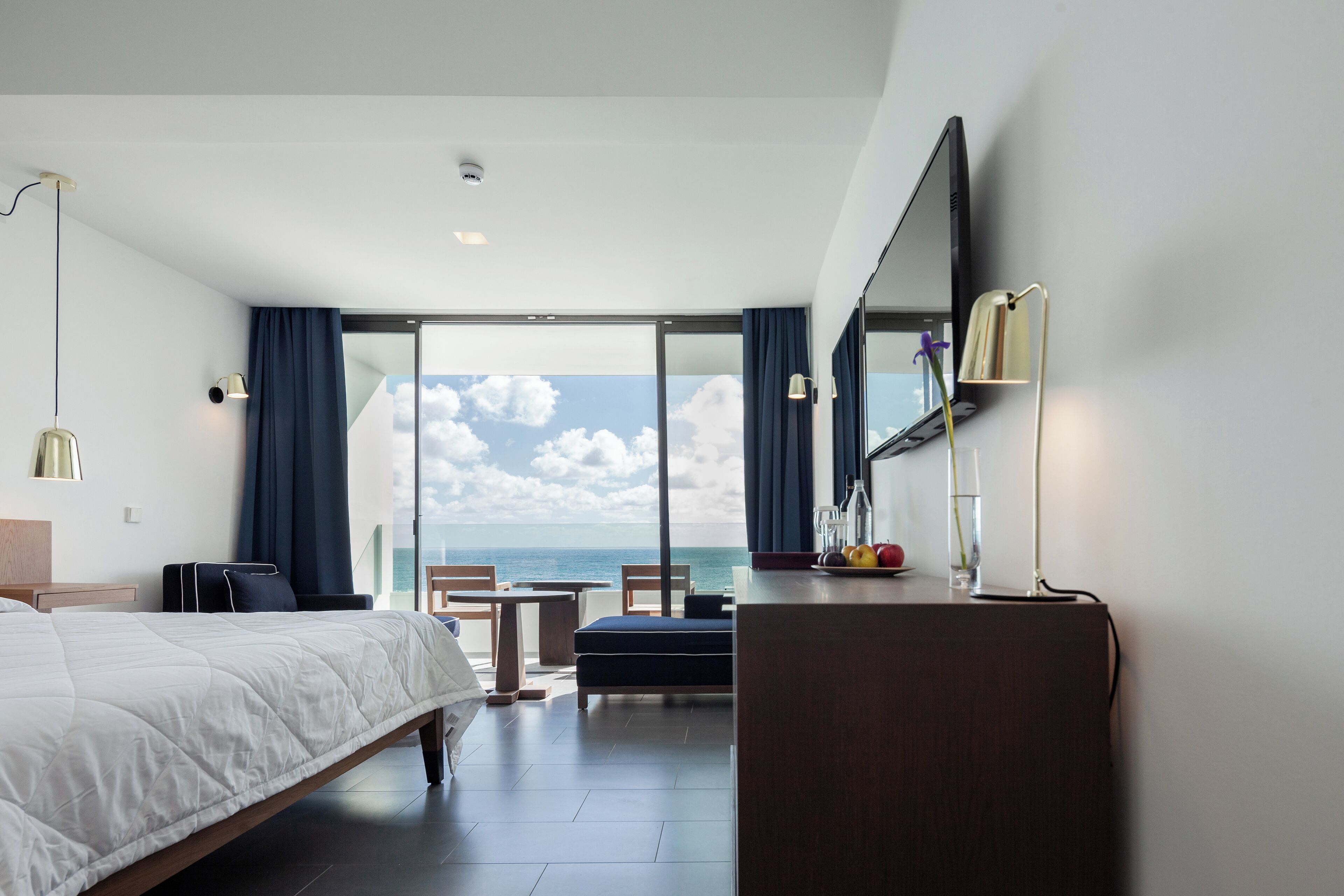 A hotel room with a bed, a desk, a chair, a table, and a large window with a view of the sea.