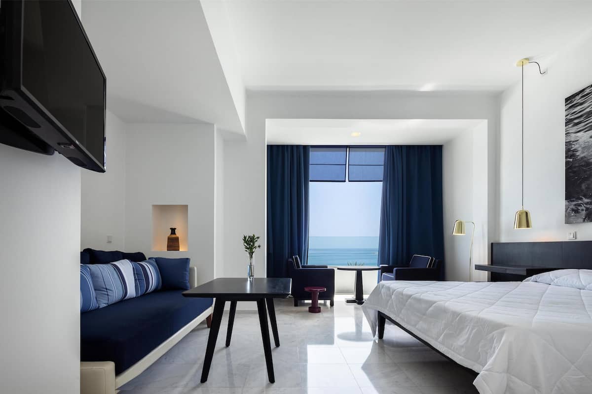 A modern hotel room with a bed, a sofa, a desk, and a view of the sea.