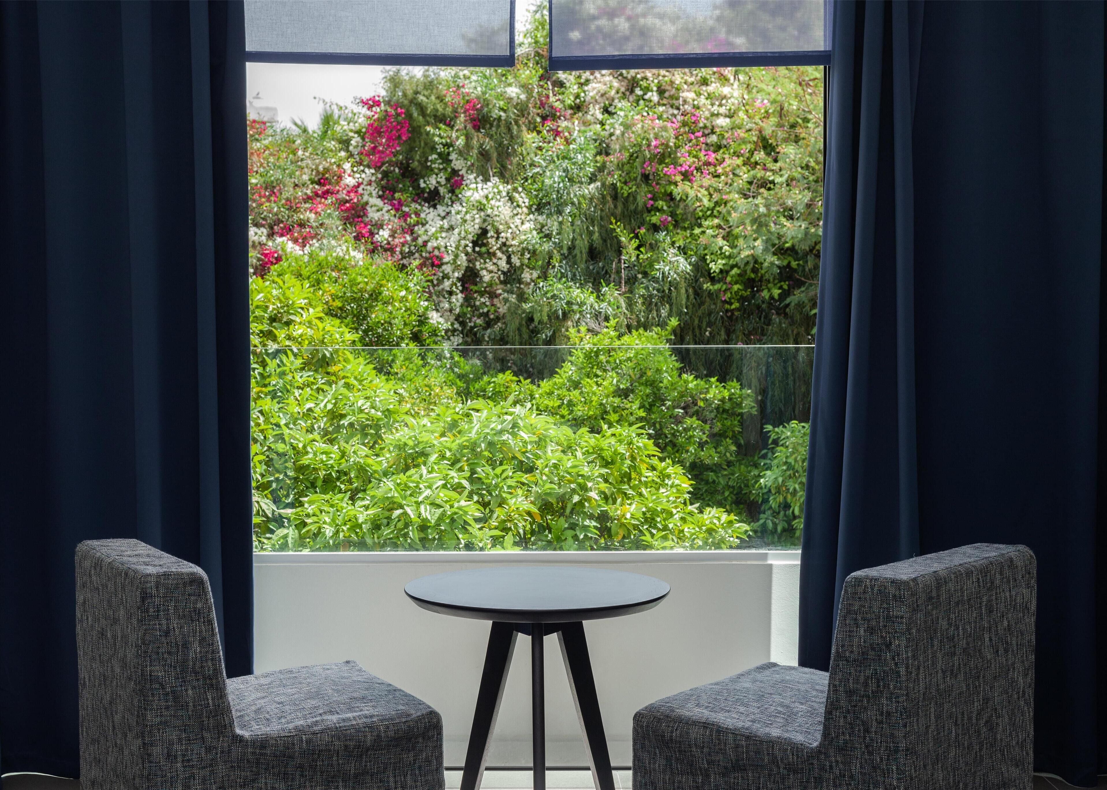 A room with a view of a garden through a window, featuring a round table and two chairs.