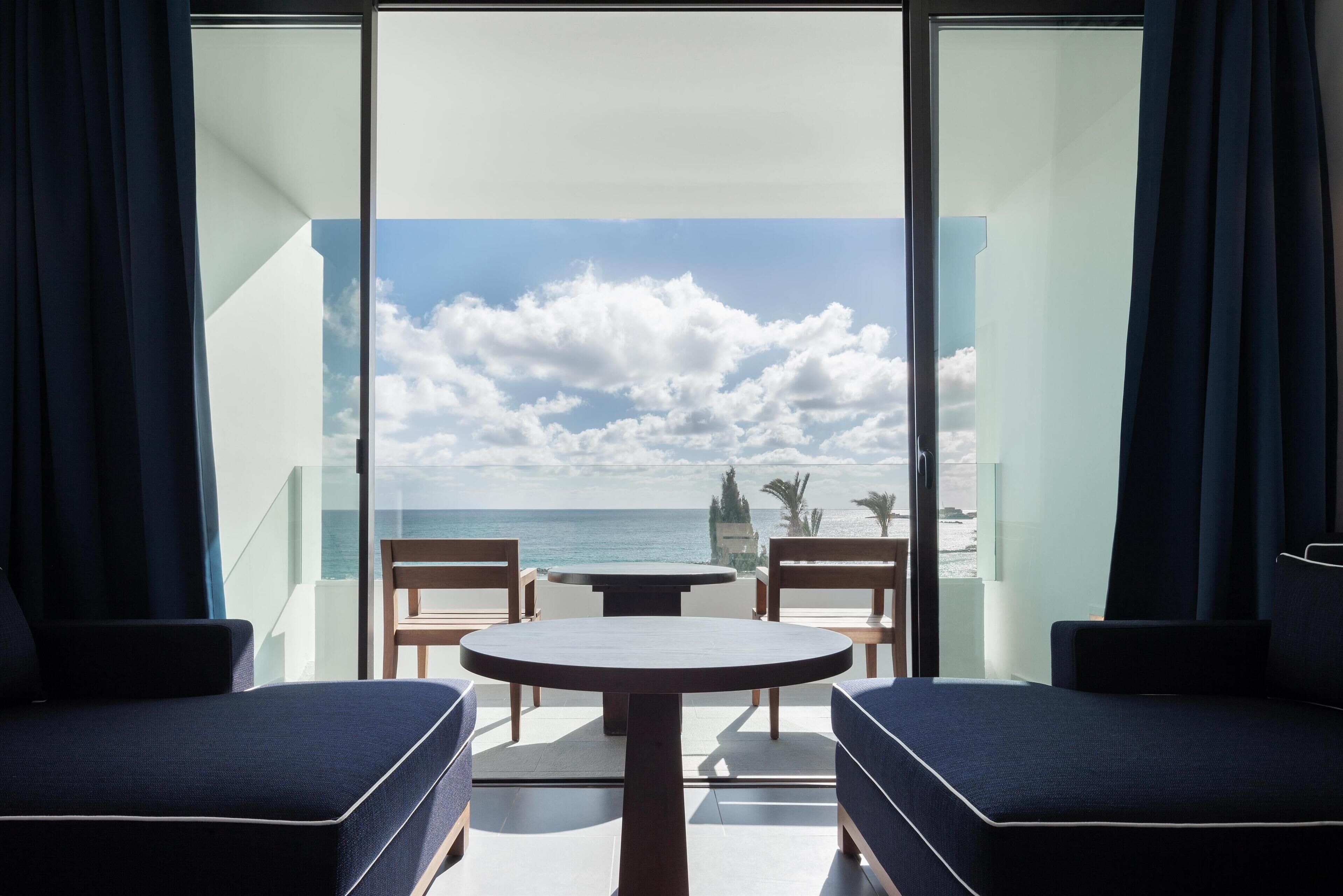 Un balcón con una mesa redonda y dos sillas azules con cojines, con vistas a una playa y un cielo despejado con nubes.