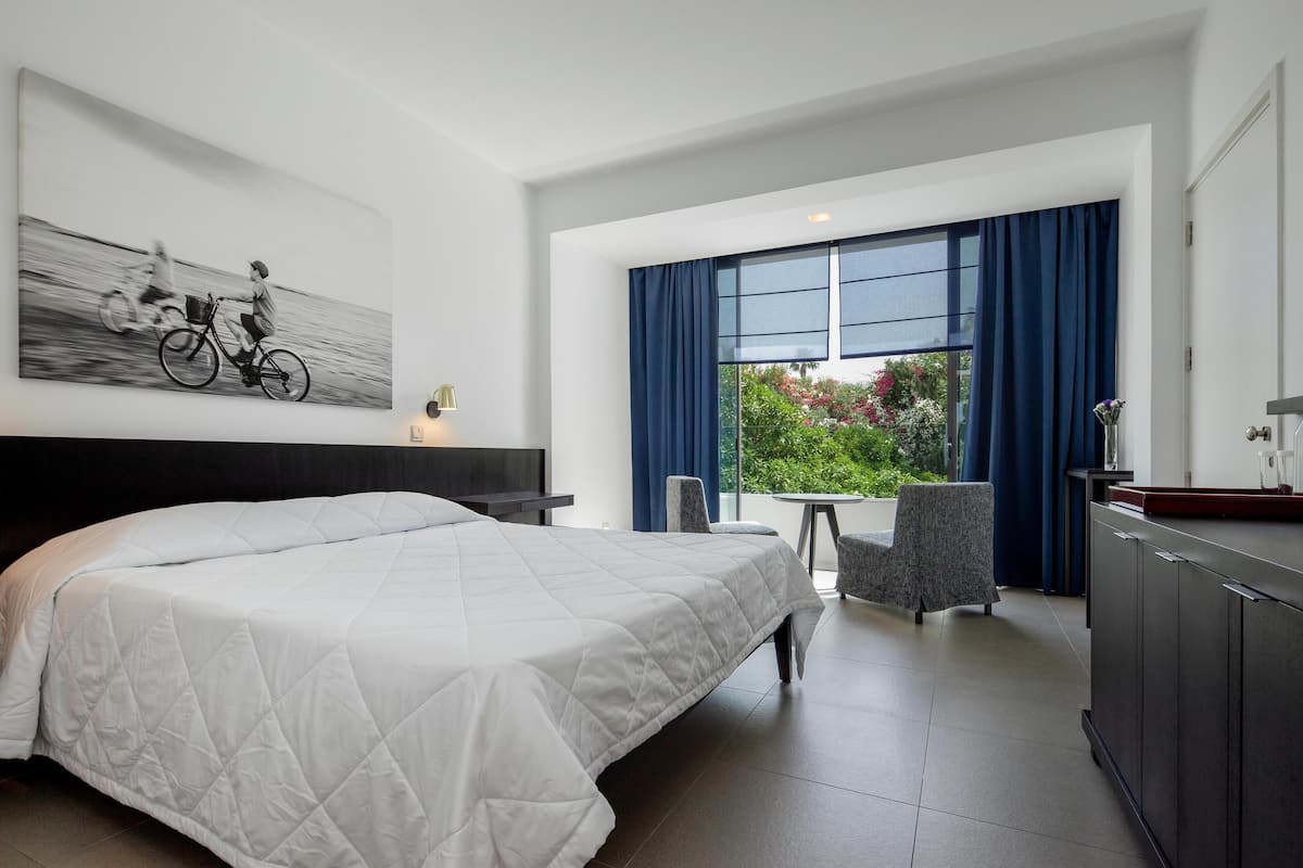 A modern hotel room with a large bed, a dining area with a table and chairs, and a view of greenery through the window.