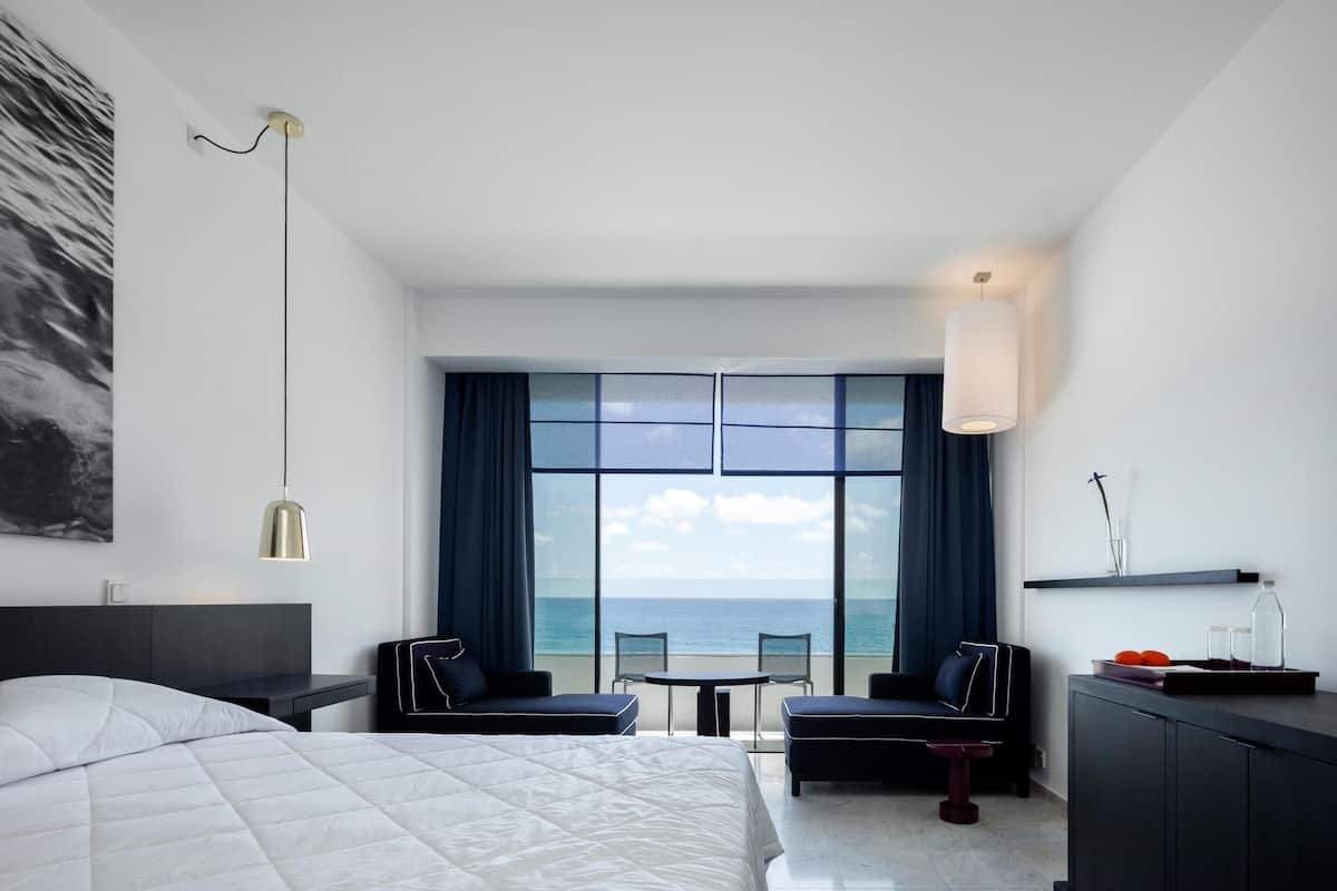 A modern hotel room with a large bed, a desk, two chairs, a view of the sea, and a large window.