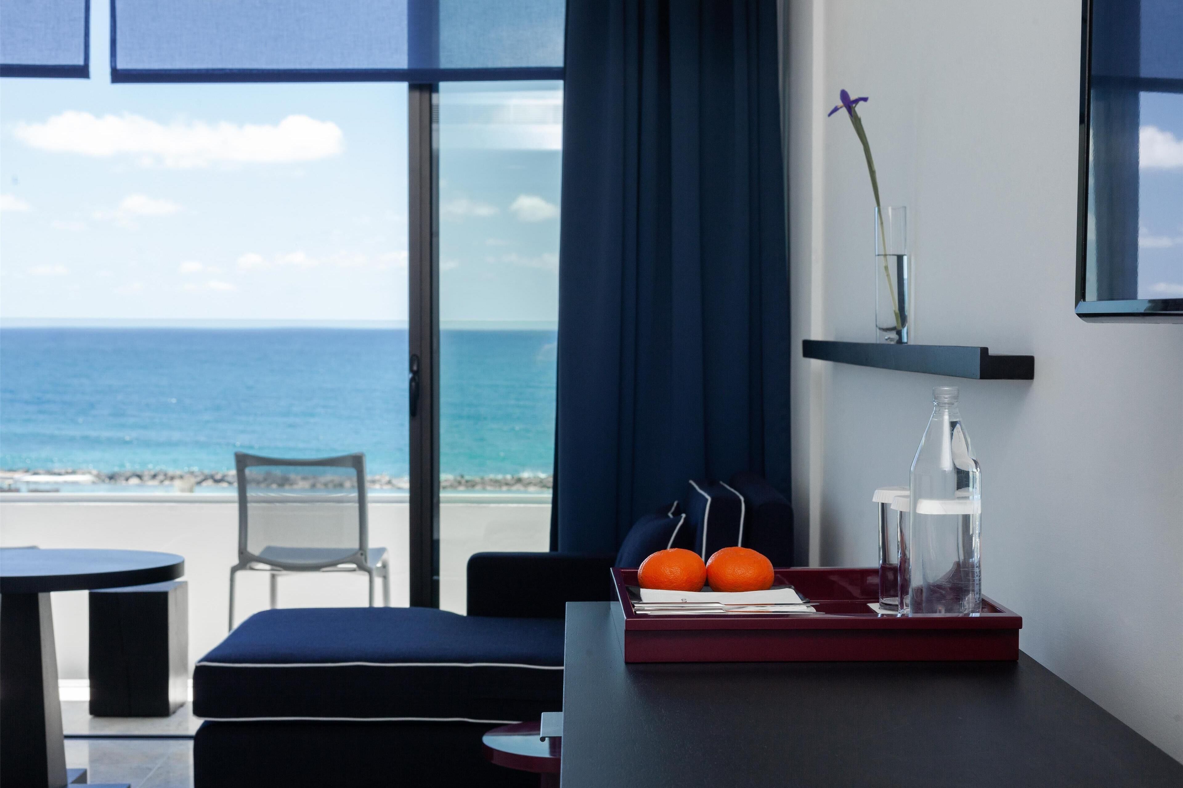 A modern hotel room with a view of the ocean, a dining table with a tray of oranges, a chair, and a small shelf with a vase.