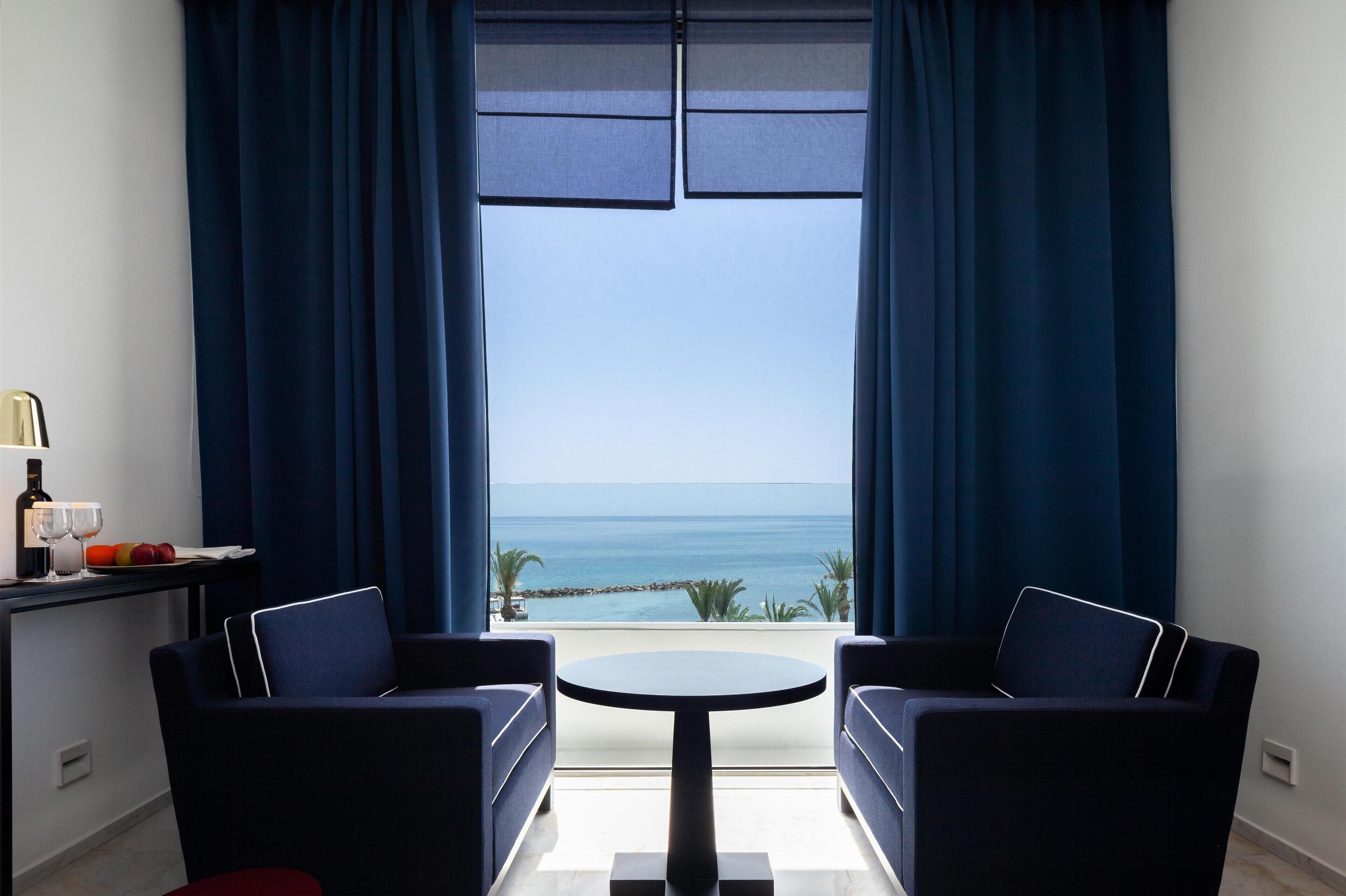 Une pièce avec vue sur la mer, deux canapés bleus, une petite table ronde et une bouteille de vin posée sur une table d’appoint.