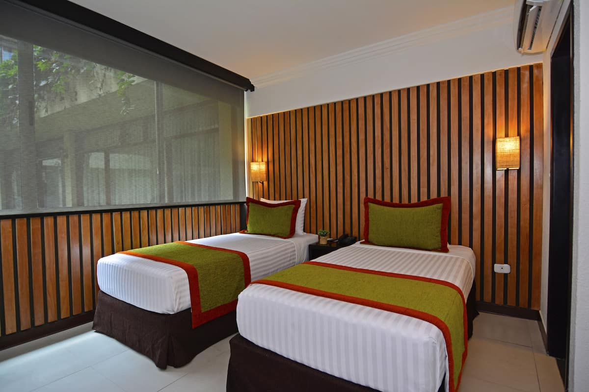Habitación de hotel con dos camas, un ventanal grande, paneles de madera en la pared y dos luces fijadas a la pared.