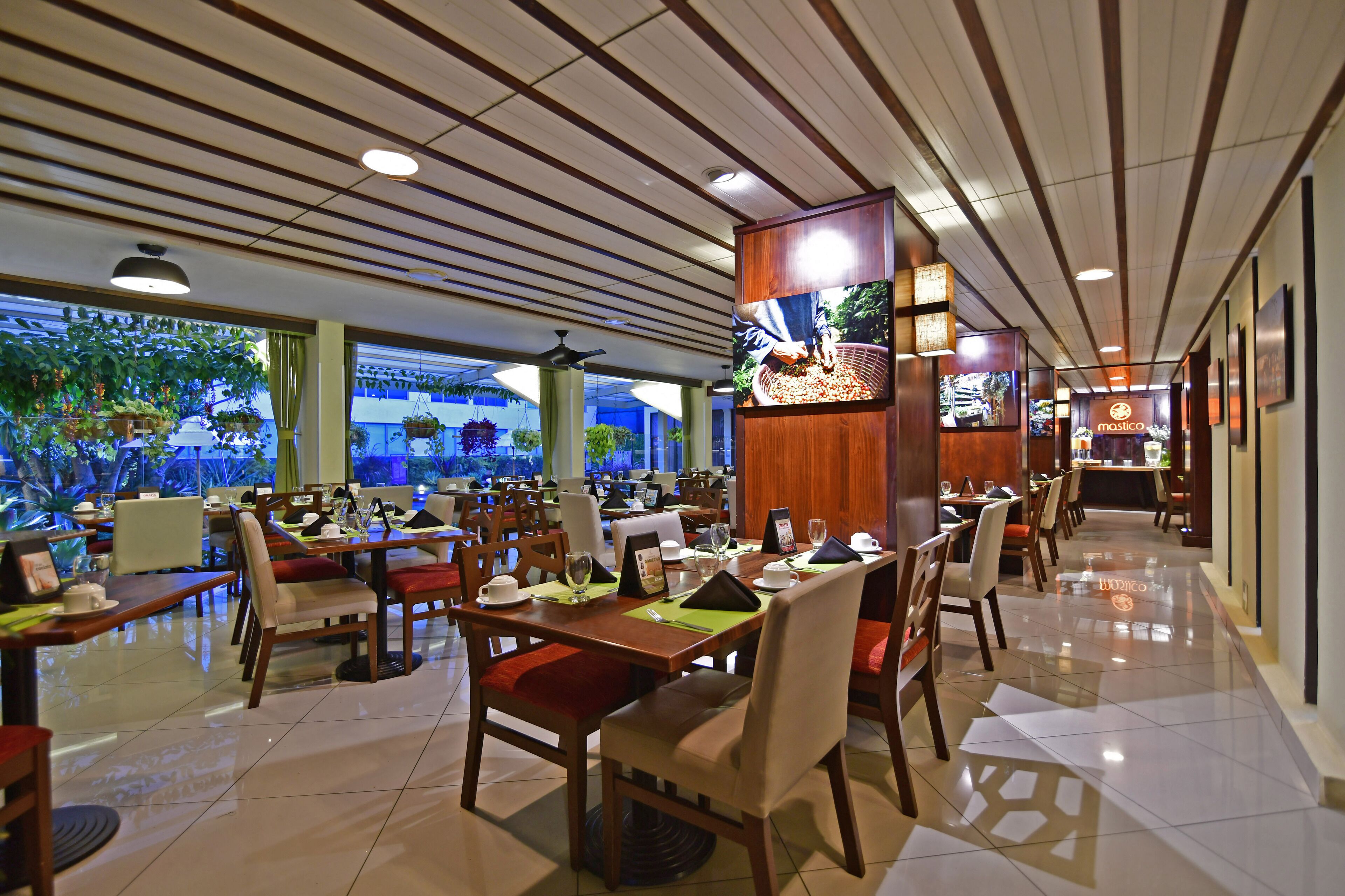 A spacious restaurant with multiple dining tables and chairs, a large TV screen, and a view of greenery outside.