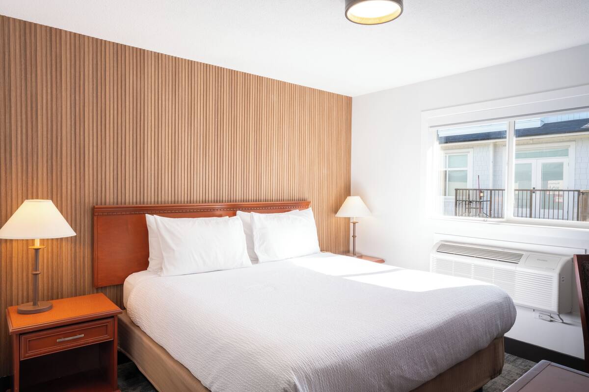Ein Hotelzimmer mit Bett, Nachttischen, Lampen, einem Fenster mit Aussicht und einer Wand mit vertikalen Linien.