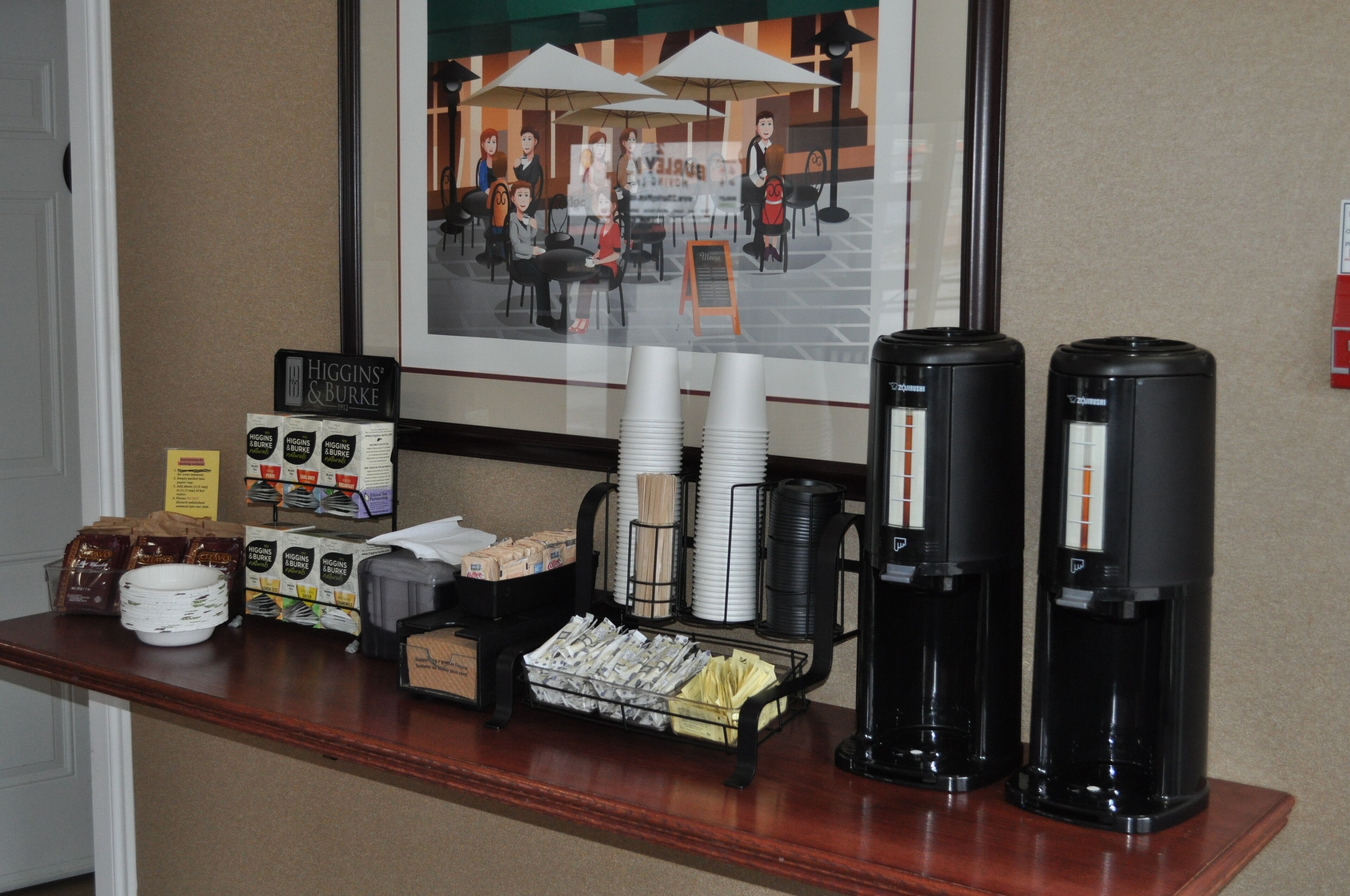 A hotel reception area with a coffee station, including coffee makers, cups, and a framed picture of a café scene.