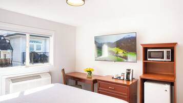 A hotel room with a bed, a desk, a chair, a small refrigerator, a microwave, a coffee maker, a vase with flowers, a painting, and a window with a view of a building and a balcony.