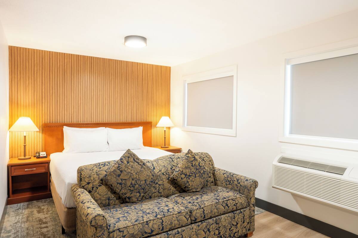 Ein Hotelzimmer mit einem Bett, zwei Nachttischlampen, einer blumigen Couch, einem hölzernen Kopfteil, einem Fenster mit Klimaanlage und einem an der Wand befestigten Bilderrahmen.