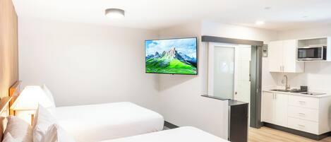 A hotel room with two beds, a TV, a kitchen area, and a door leading to the bathroom.