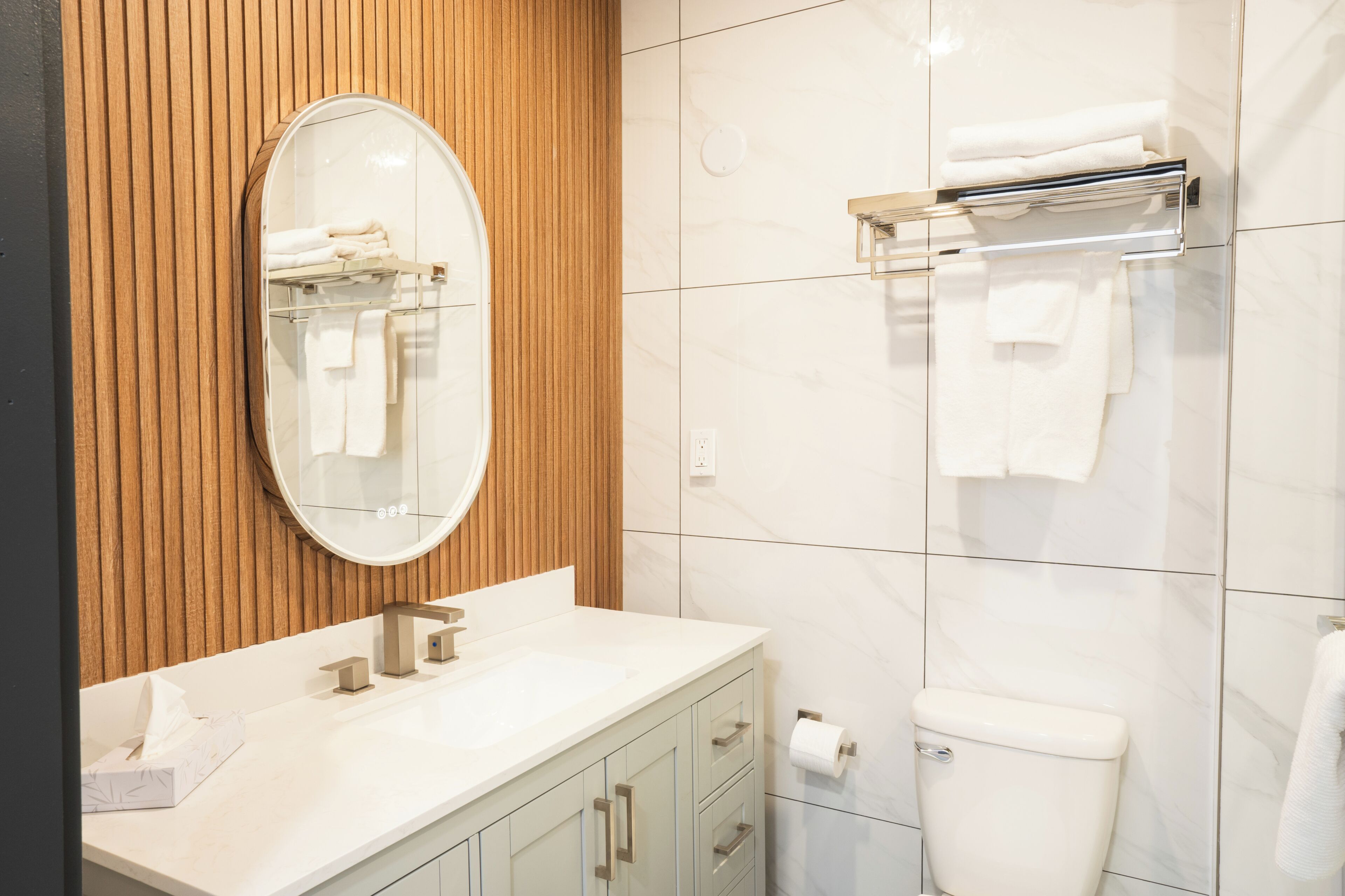 A modern bathroom with a wooden panel wall, a round mirror, a white sink, a toilet, and a towel rack.