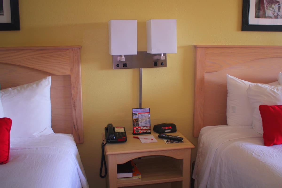 A hotel room with two beds, each with white bedding and red pillows. There is a bedside table with a phone, a remote control, and a menu card.