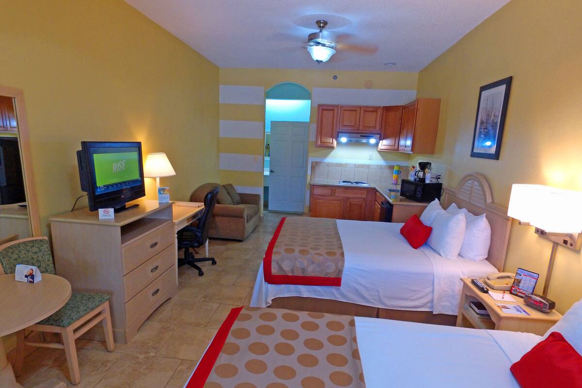 A hotel room with a bed, a desk with a computer, a chair, a small table, and a kitchenette.