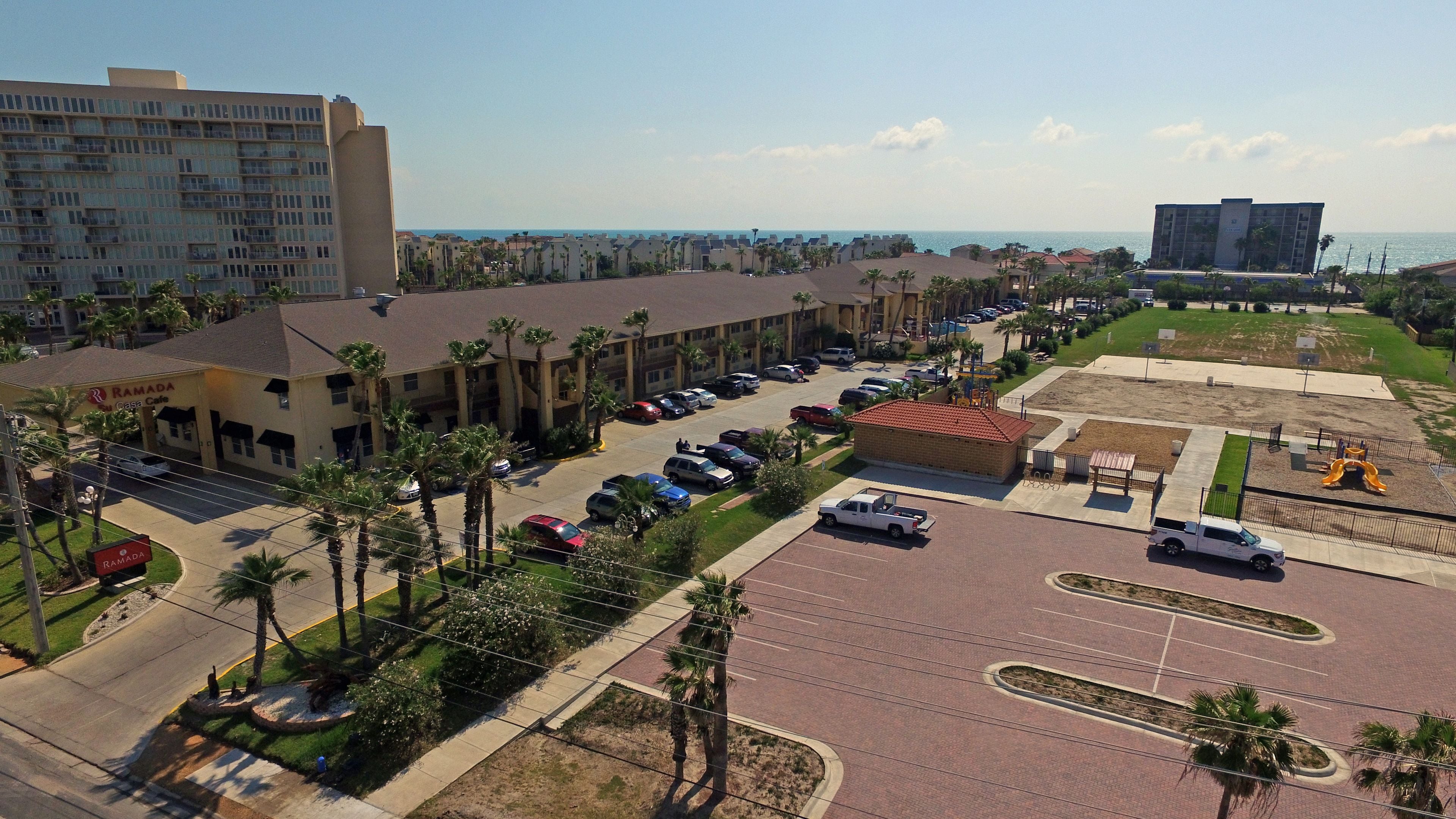 A parking lot with multiple cars, a building with a sign reading "RAMADA," and a view of the ocean in the background.