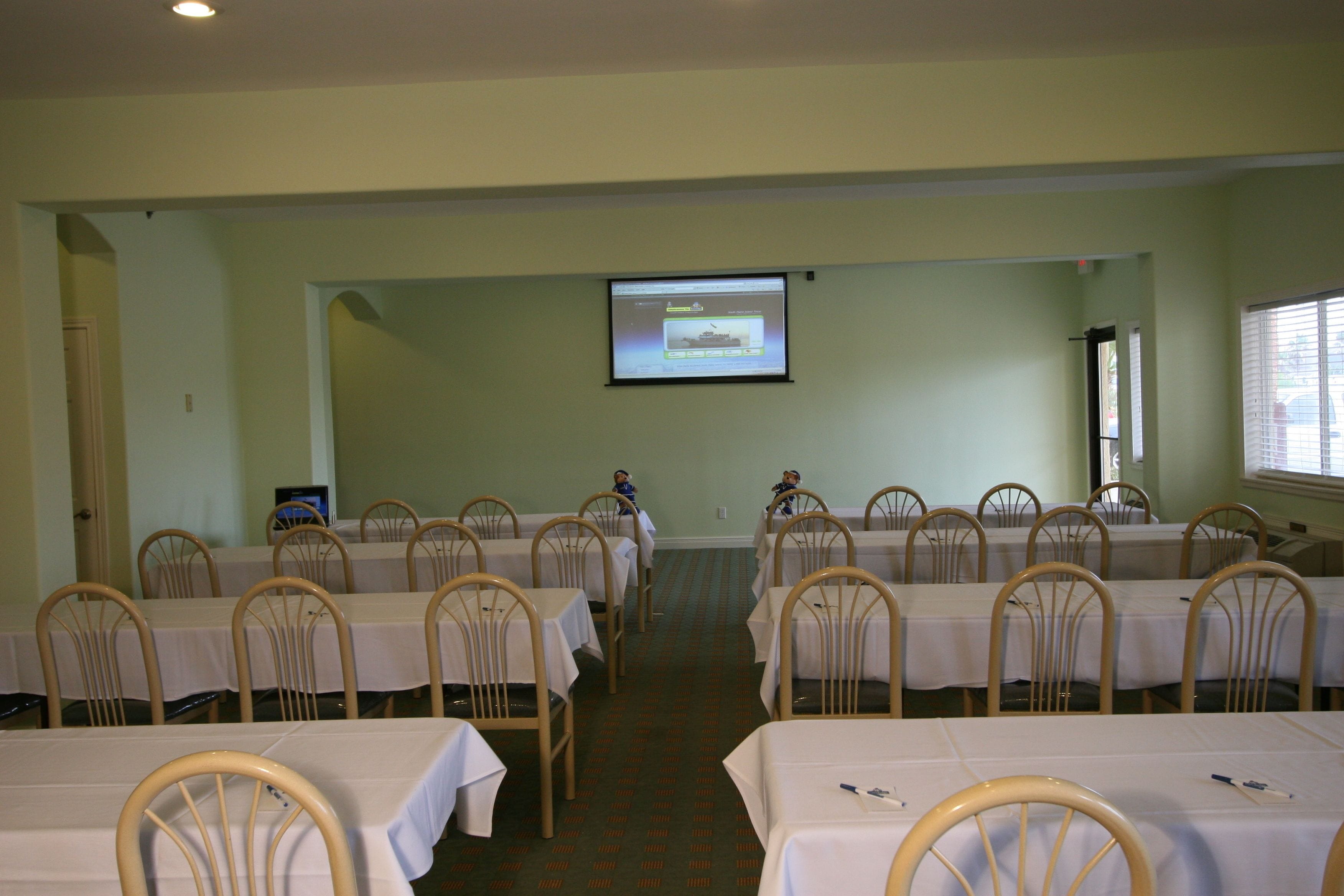 Un salón de conferencias con filas de mesas y sillas, una pantalla de proyector al frente y una ventana a la derecha.