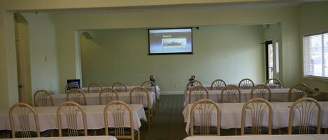 Un salón de conferencias con filas de mesas y sillas, una pantalla de proyector al frente y una ventana a la derecha.