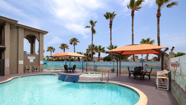 A swimming pool with lounge chairs and umbrellas, surrounded by palm trees and a mural of a beach scene. - Ramada by Wyndham & Suites South Padre Island At the Beach (South Padre Island)
