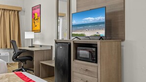 A hotel room with a bed, a desk with a chair, a refrigerator, a microwave, a TV displaying a beach scene, and a poster on the wall. - Ramada by Wyndham & Suites South Padre Island At the Beach (South Padre Island)