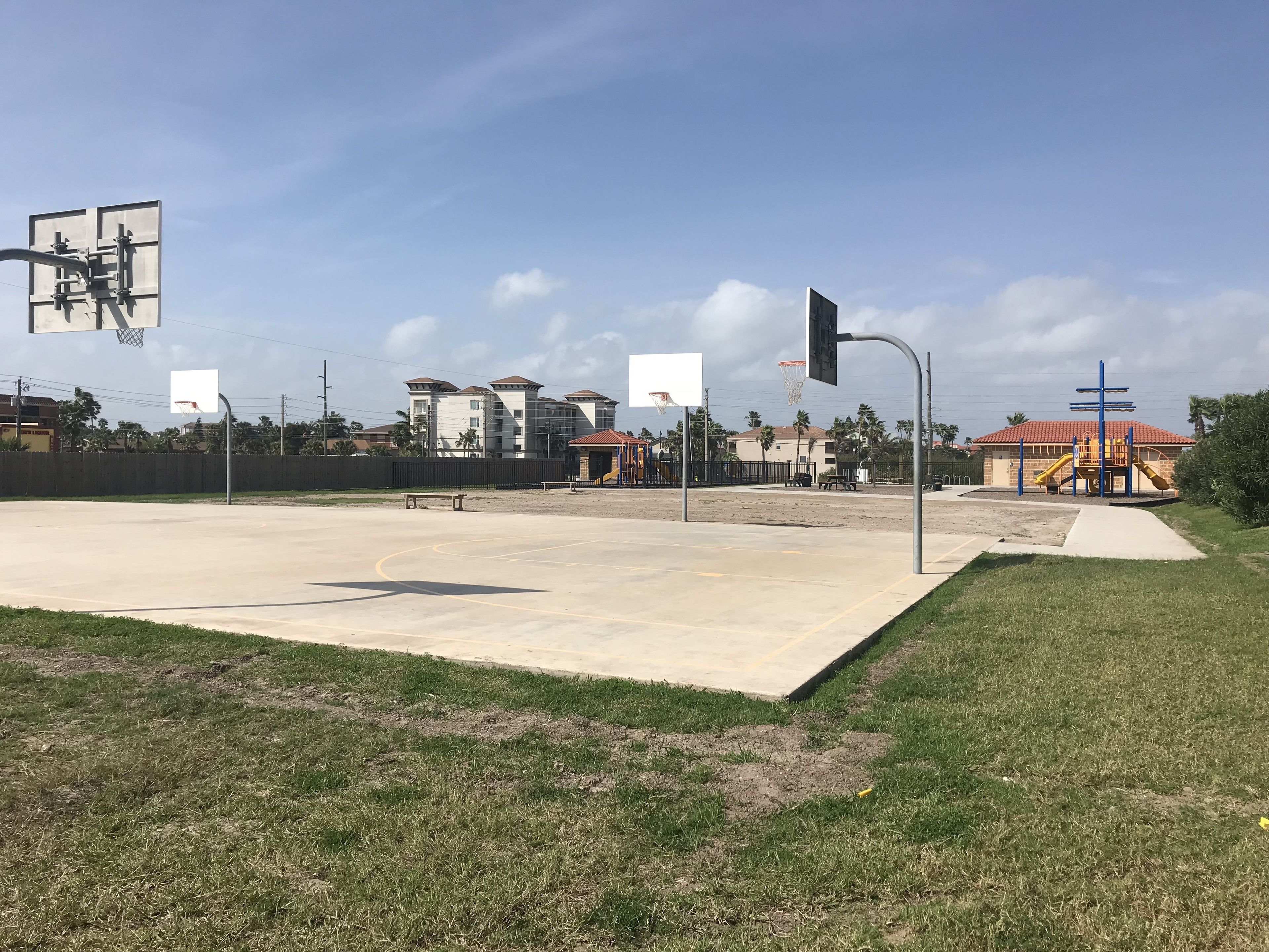 An outdoor basketball court with a fence, a playground, and houses in the background.