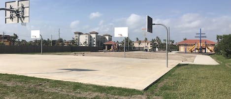 An outdoor basketball court with a fence, a playground, and houses in the background.
