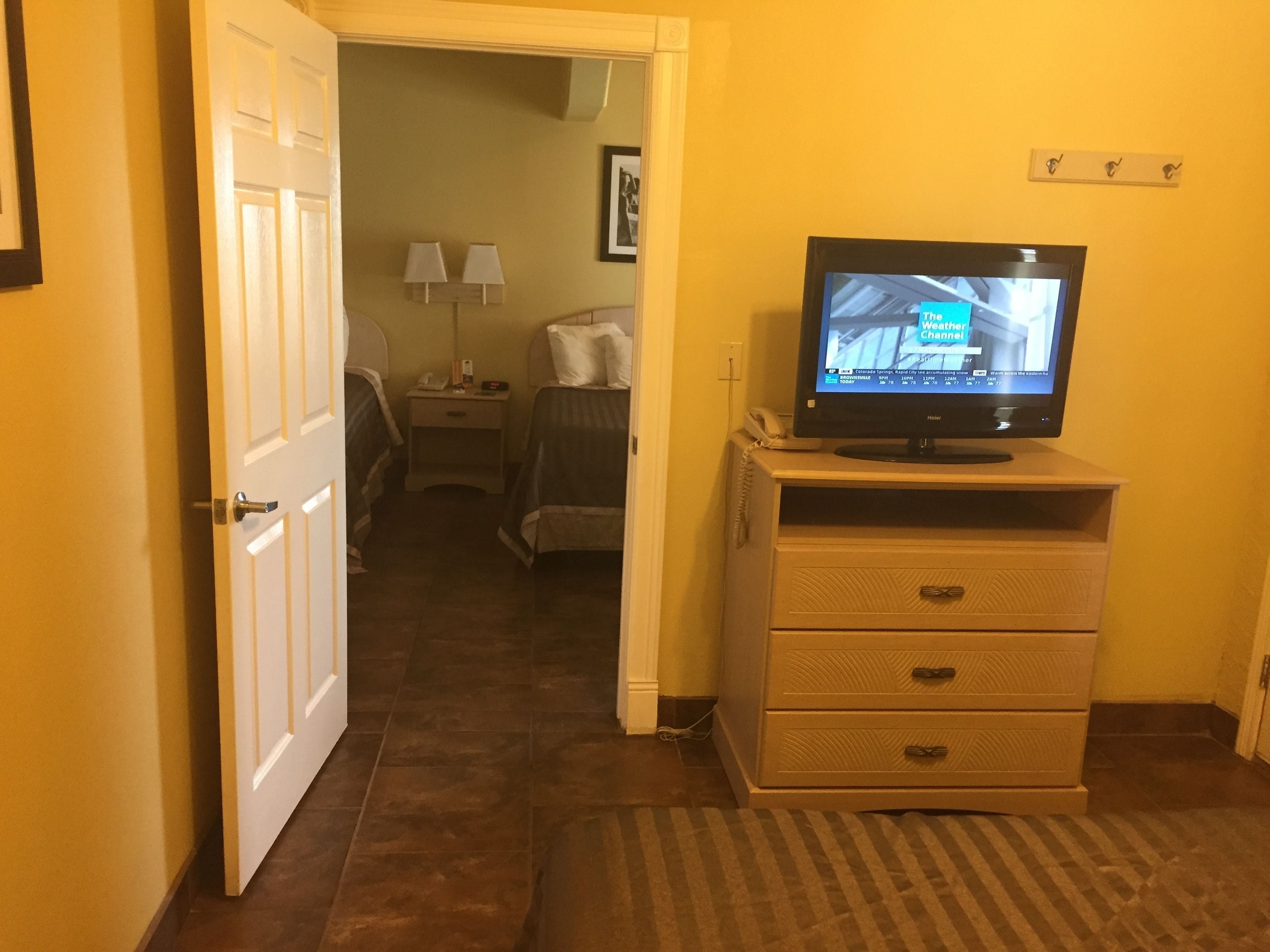 Une chambre d’hôtel avec une télévision posée sur une commode, un lit, une table de chevet avec des lampes et une porte donnant sur une autre pièce.