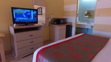 Habitación de hotel con televisor mostrando un pronóstico del tiempo, cama con colcha estampada, microondas, pequeño mueble y zona de lavamanos.
