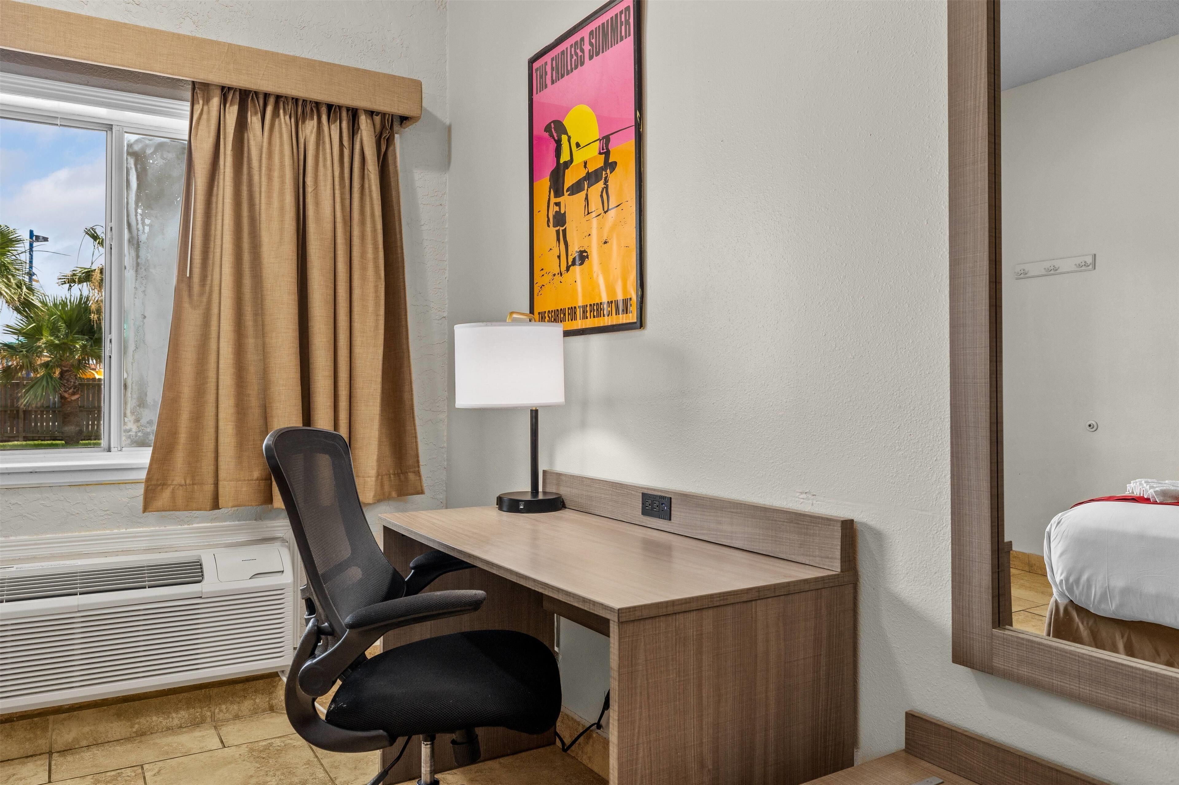 A hotel room with a desk, chair, and a poster on the wall.