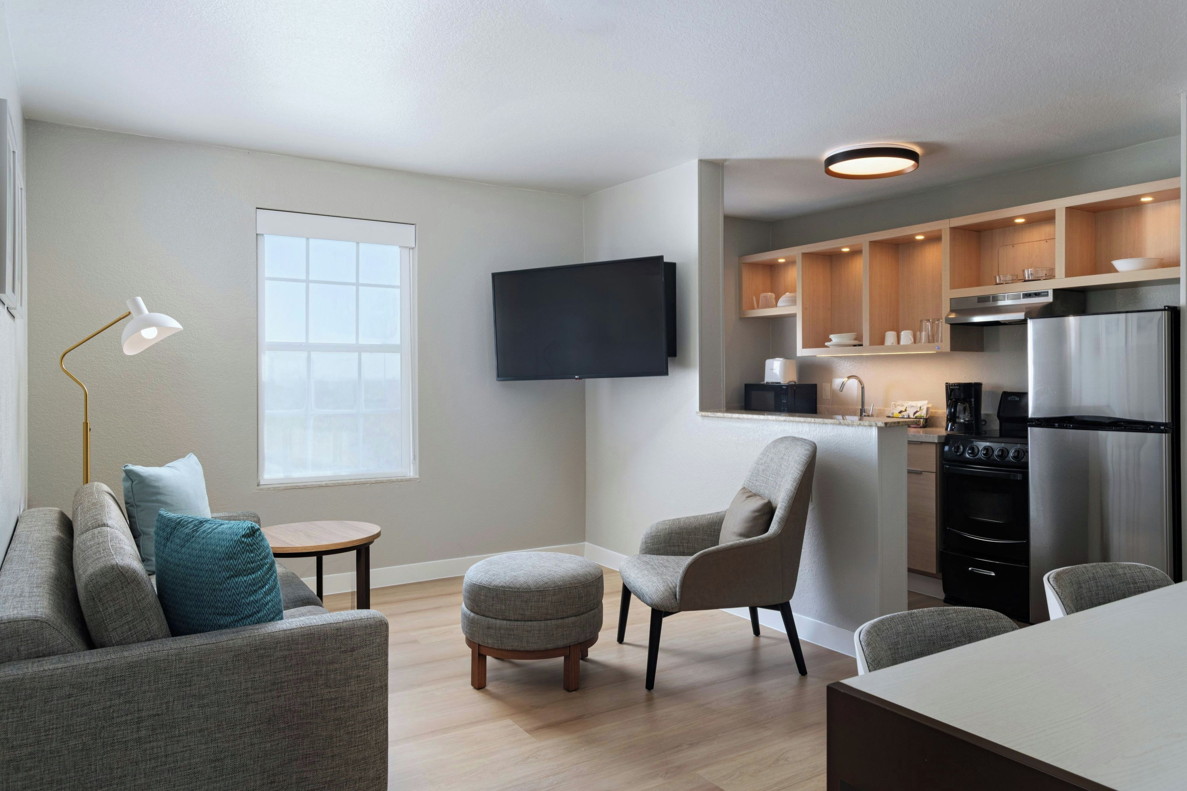 A modern living room with a sofa, armchair, and a small round table. A flat-screen TV is mounted on the wall, and a ceiling light is visible.