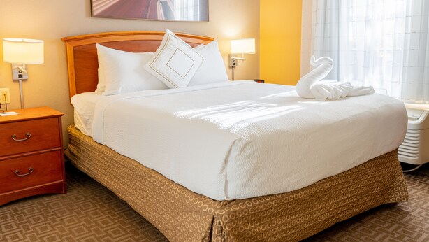A neatly made bed with a wooden headboard, two bedside lamps, and a swan-shaped towel on the bed. - TownePlace Suites by Marriott Dallas Las Colinas (Irving)