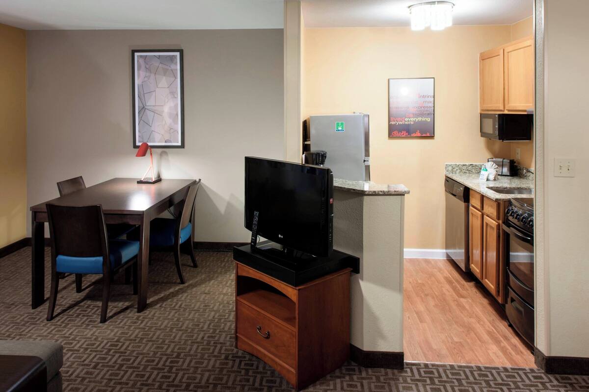 A compact hotel room with a dining area, kitchenette, and a flat-screen TV on a wooden stand.