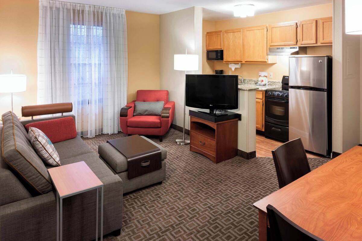 A hotel room with a living area, a sofa, a red armchair, a TV, a kitchen with wooden cabinets, a refrigerator, and a patterned carpet.