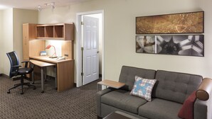 A hotel room with a wooden desk, a grey sofa, a patterned carpet, and a door. - TownePlace Suites by Marriott Dallas Las Colinas (Irving)