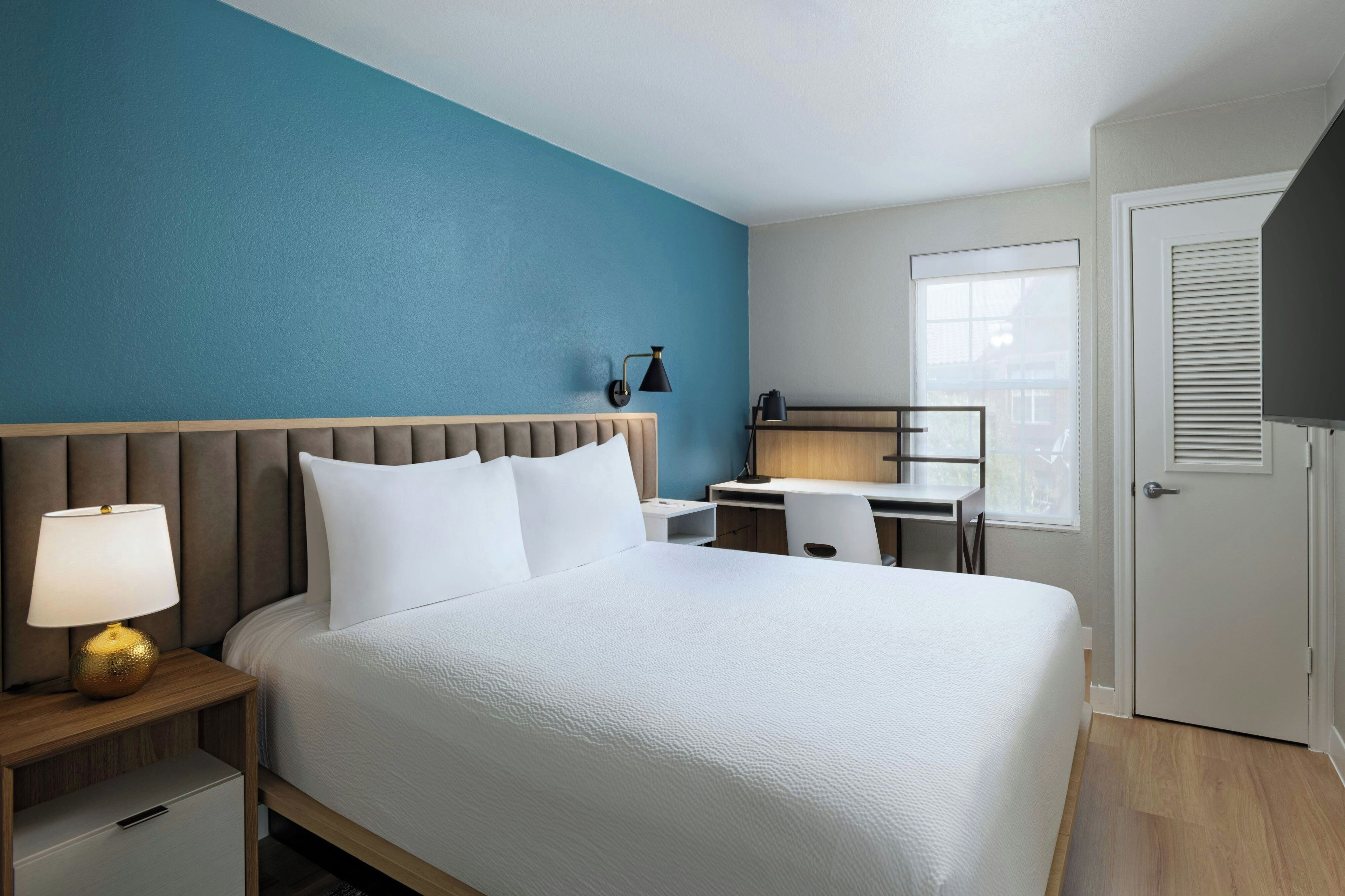 A hotel room with a bed, bedside table, lamp, desk, chair, and a door.