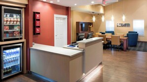 A hotel reception area with a counter, a refrigerator stocked with beverages, and a seating area with tables and chairs. - TownePlace Suites by Marriott Dallas Las Colinas (Irving)