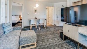 A modern hotel room with a grey sofa, a flat-screen TV, a dining area with two chairs, and a small kitchen area.