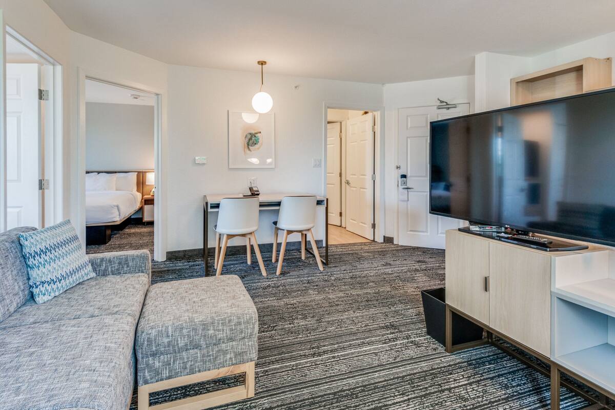 Una camera d'albergo moderna con un divano grigio, una televisione a schermo piatto, un'area pranzo con due sedie e un piccolo angolo cottura.