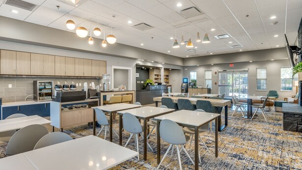 A modern dining area with tables and chairs, a kitchen area with appliances, and a patterned carpet.